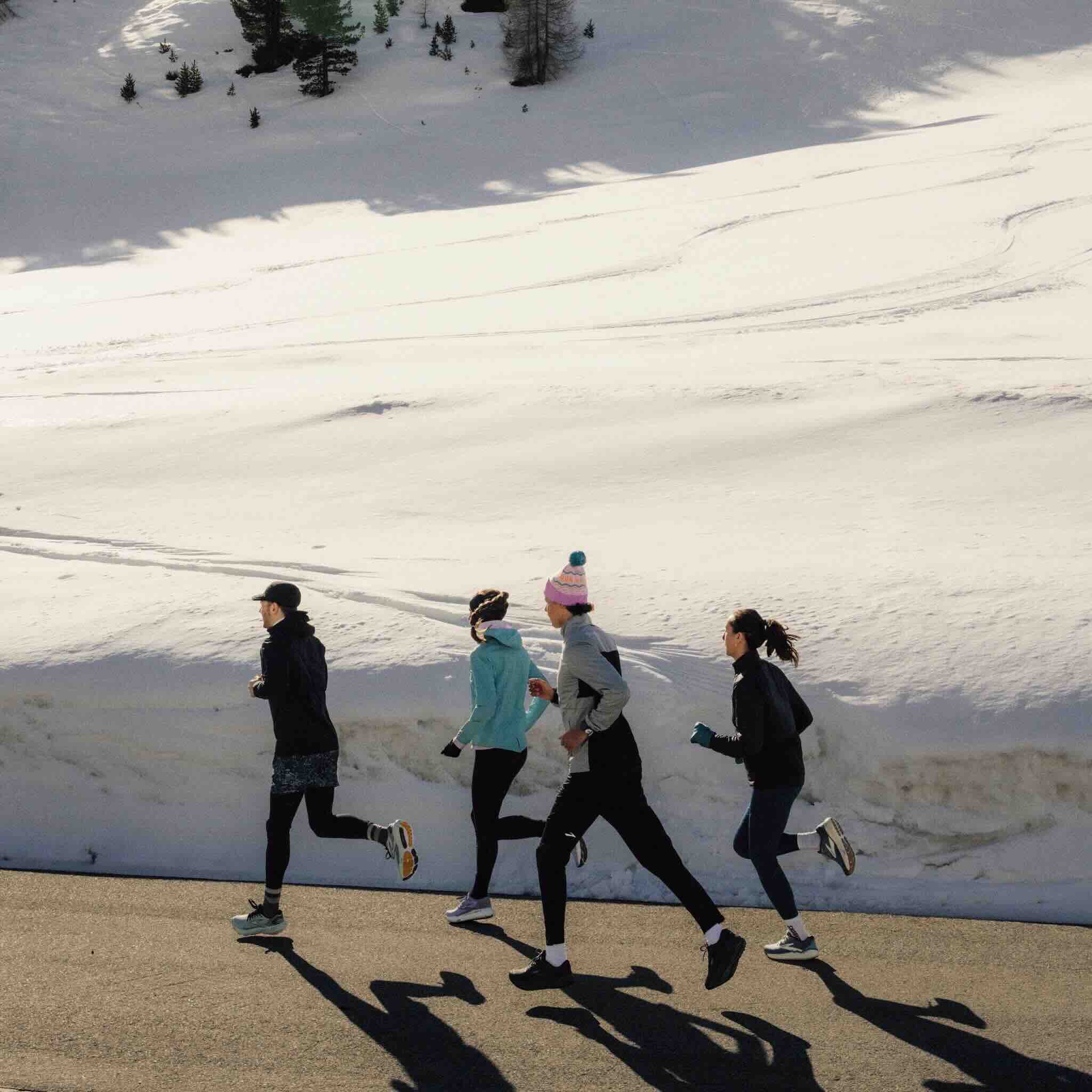 Four runners jogging in single file along a path with a snowy mountain slope in the background. The group includes one man in front wearing a black jacket and shorts, followed by three women in winter running gear including tights, jackets, and winter hats/headbands. The scene appears to be shot during a winter morning or afternoon, with strong shadows cast on the path. Behind them, a pristine snow-covered slope rises up with scattered evergreen trees. The runners appear to be carrying water bottles or running gear, suggesting a group training run or social exercise session