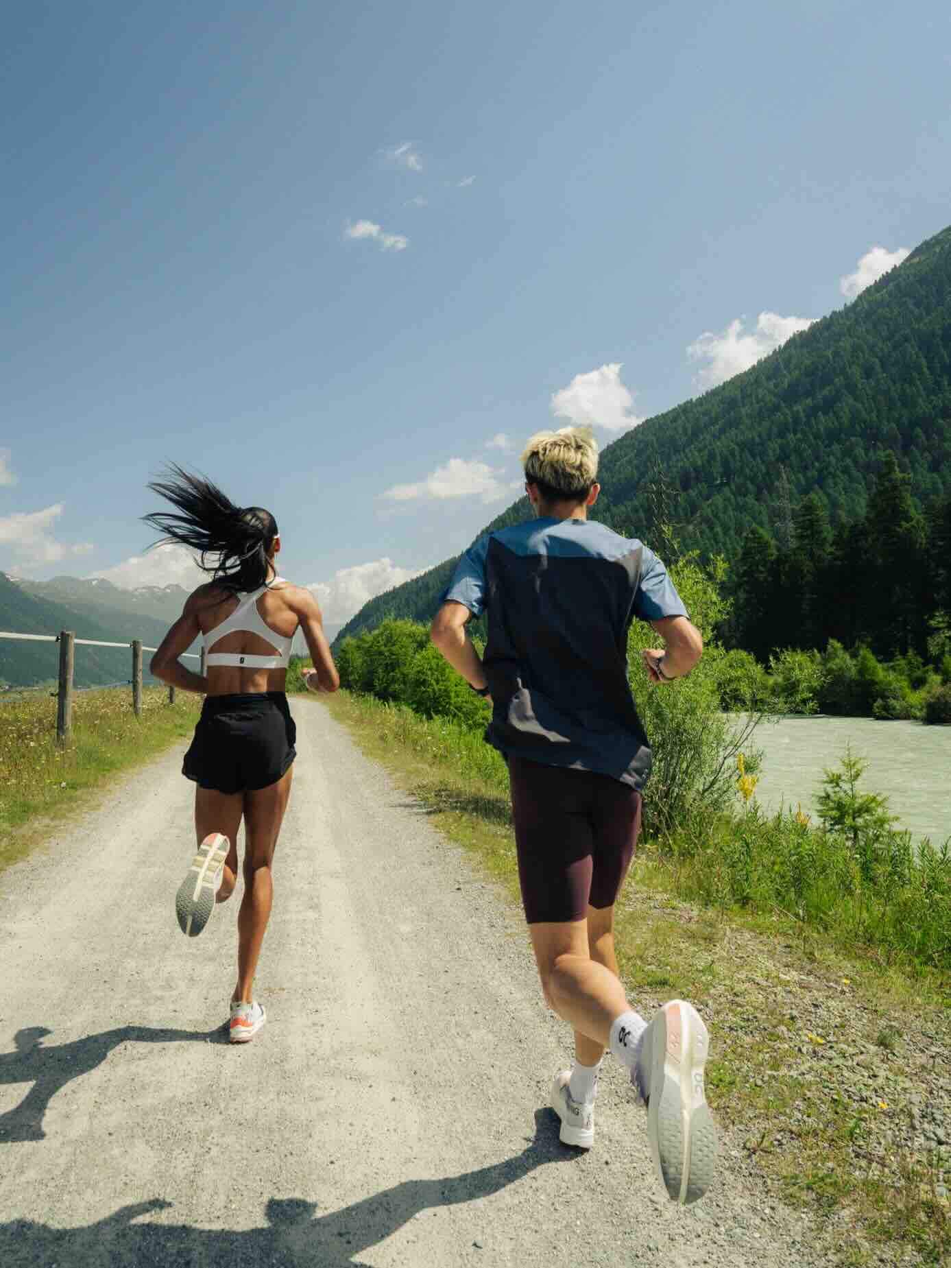 Two runners on a mountain trail on a sunny day. A woman in a white sports bra and black shorts leads, her long dark hair streaming behind her, while a man in a blue t-shirt and black running shorts follows. They're running on a dirt path bordered by wooden fence posts and wildflowers, with a pine-covered mountain slope and turquoise river visible in the background. The lighting suggests mid-day, with bright blue skies and scattered clouds. Both runners are wearing white athletic shoes and appear to be captured mid-stride in their run.