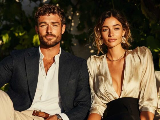 A sophisticated outdoor lifestyle photograph featuring a fashionable couple dressed elegantly for a formal evening gathering. The man wears a tailored navy blazer over a crisp white button-down shirt, paired with beige trousers and a brown leather belt, exuding timeless refinement. The woman stuns in a champagne-colored satin blouse with a plunging neckline and voluminous sleeves, tucked into high-waisted black trousers. She accessorizes with gold statement earrings and a delicate chain necklace, while her hair is styled in loose, romantic waves. They pose confidently in a sun-drenched setting with lush greenery in the background, seated at a table with glasses of white wine, capturing the essence of an elegant, upscale soirée.